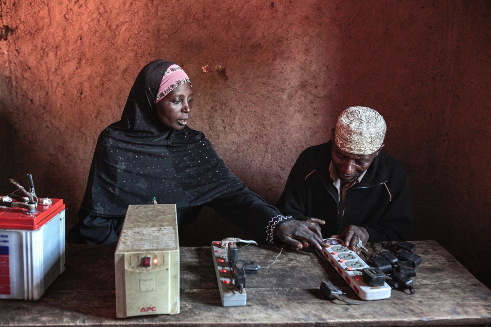 Maida Mushimiyimana | Mobile Phone Charging | Duteranink Unga, Rwanda. Man and woman sitting at a table with two batteries, electrical sockets and phone chargers.