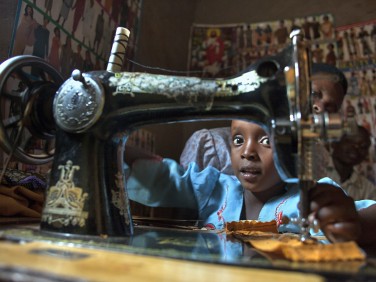 Student of Odette Mukarusagara | Tailor | Kiziguro Gatsibo, Rwanda. Young girl looking at camera behind a sewing machine.