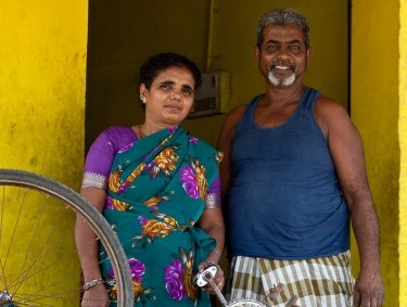 Najma standing outside a yellow house with a man and a bicycle that is being repaired.